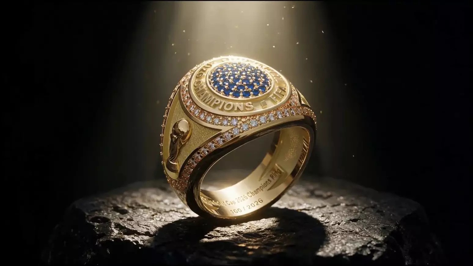 Anel dourado de campeão da Copa do Mundo FIFA, com pedras azuis no centro e diamantes ao redor, sobre uma superfície escura e texturizada, sob um feixe de luz que destaca seu brilho