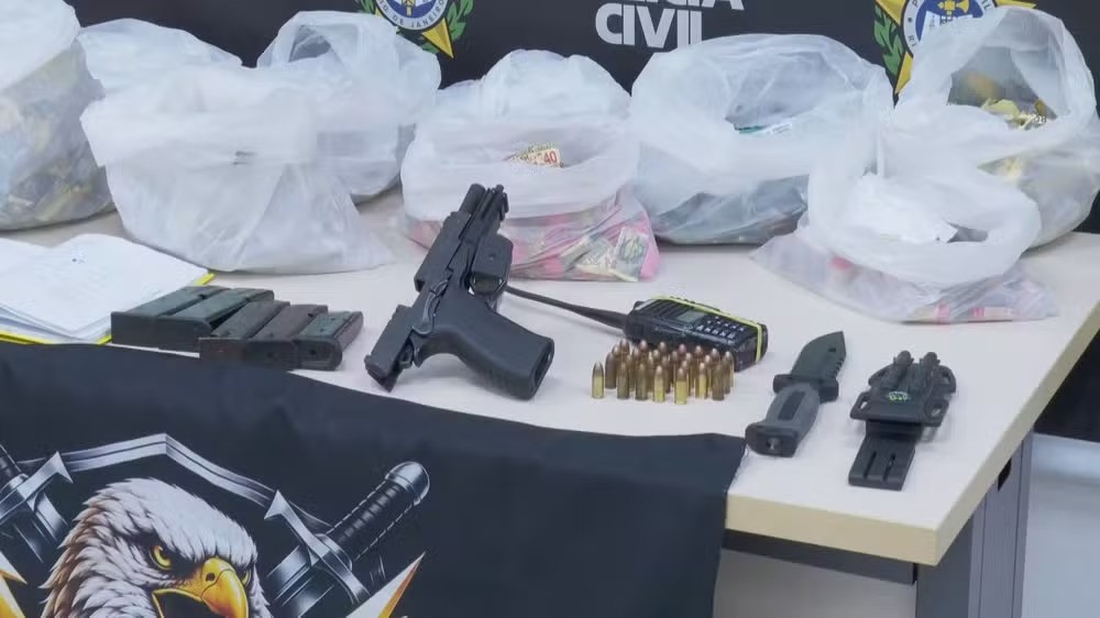 Mesa com itens apreendidos: uma pistola preta, carregadores, munições, um rádio comunicador, uma faca tática e sacolas plásticas cheias de objetos não identificados. Uma camiseta preta com estampa de águia e espadas está dobrada na frente