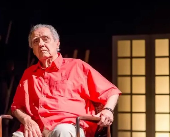 Homem idoso de cabelos grisalhos, camisa vermelha e calça clara, sentado em uma cadeira de madeira, com expressão séria, olhando para frente. Ao fundo, uma janela iluminada com grades escuras e um ambiente escuro
