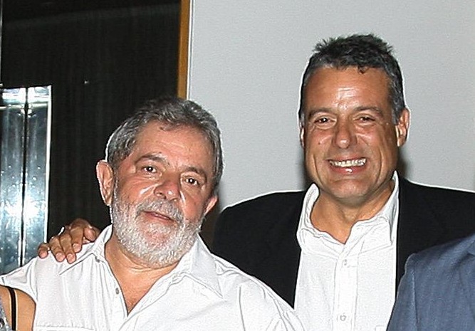 Luiz Inácio Lula da Silva, com barba grisalha e camisa branca, sorri ao lado de um homem de cabelo escuro e blazer preto, que também sorri, com a mão no ombro de Lula