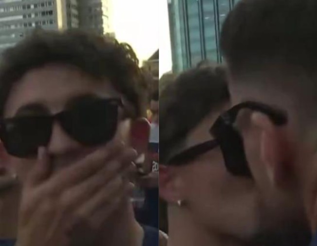 Jovem com cabelo escuro e óculos de sol escuros, cobrindo a boca com a mão direita, em um ambiente externo com prédios ao fundo