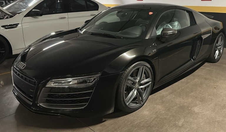 Carro esportivo Audi R8 preto brilhante estacionado em uma garagem, com um veículo branco parcialmente visível ao fundo