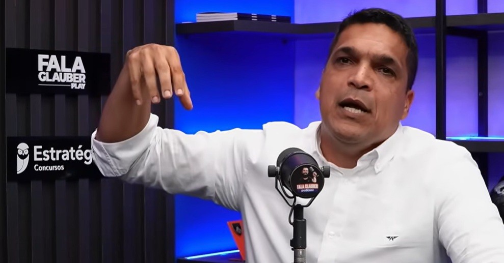 Homem de pele morena, camisa branca, gesticula com a mão direita apontando para baixo, em um estúdio com iluminação azul e placas pretas com os dizeres FALA GLAUBER PLAY e Estratégia Concursos