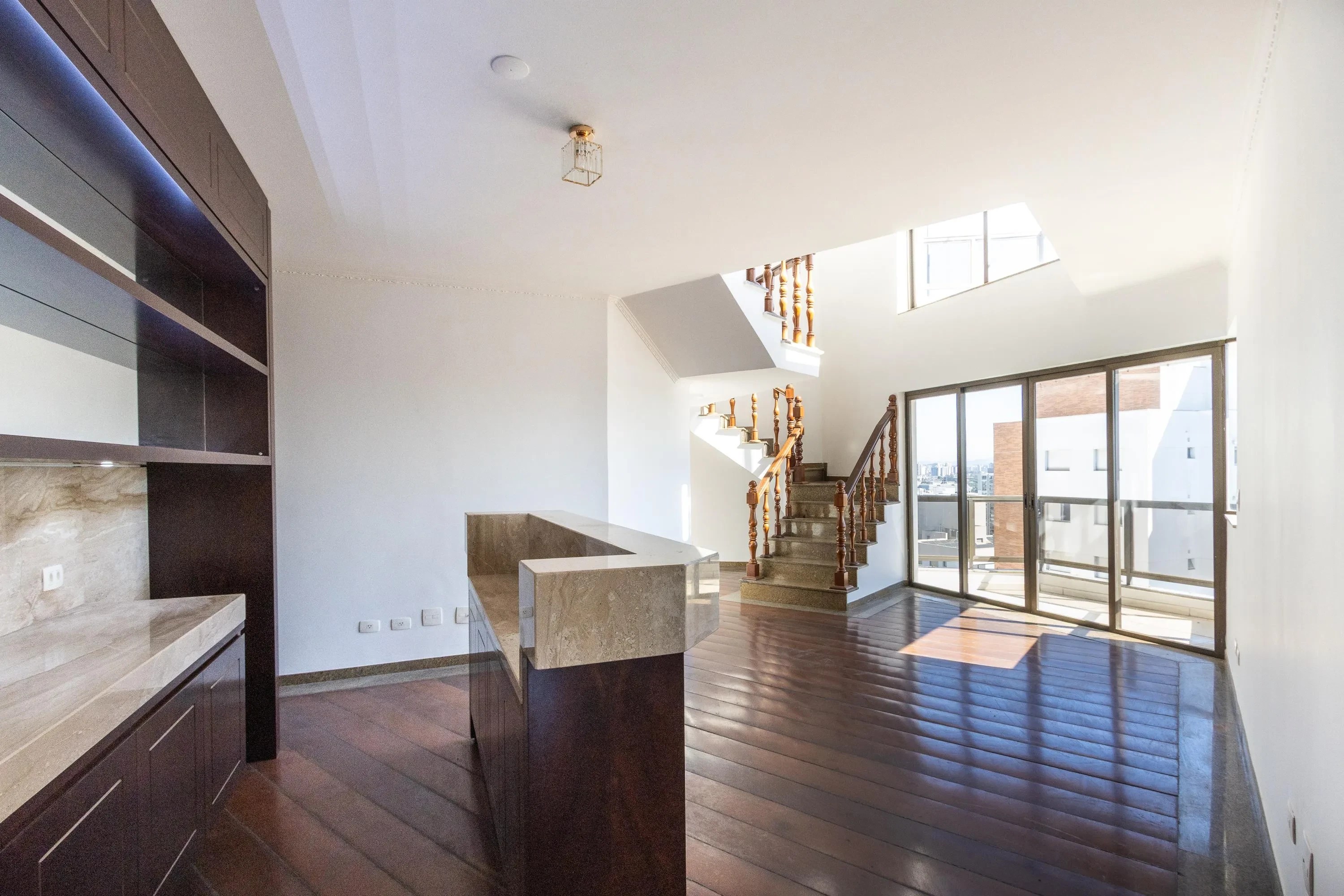 Sala ampla e iluminada com piso de madeira escura, paredes brancas e uma escada de madeira com corrimão trabalhado. À esquerda, um balcão de mármore claro e armários escuros. Ao fundo, uma porta de vidro que dá para uma varanda com vista para a cidade