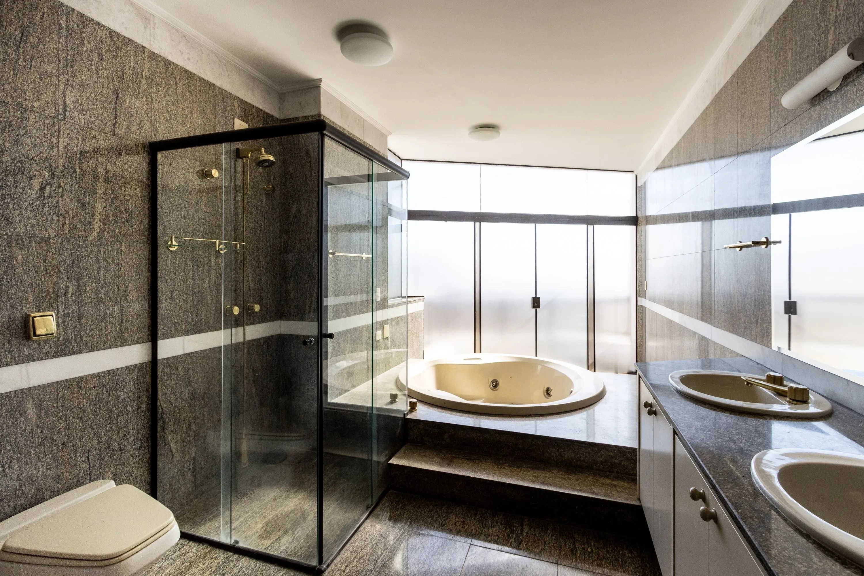 Banheiro moderno com paredes de granito cinza escuro e faixa branca, box de vidro com chuveiro dourado, vaso sanitário bege, banheira de hidromassagem elevada e duas pias ovais em bancada de granito, com espelho e janelas foscas ao fundo