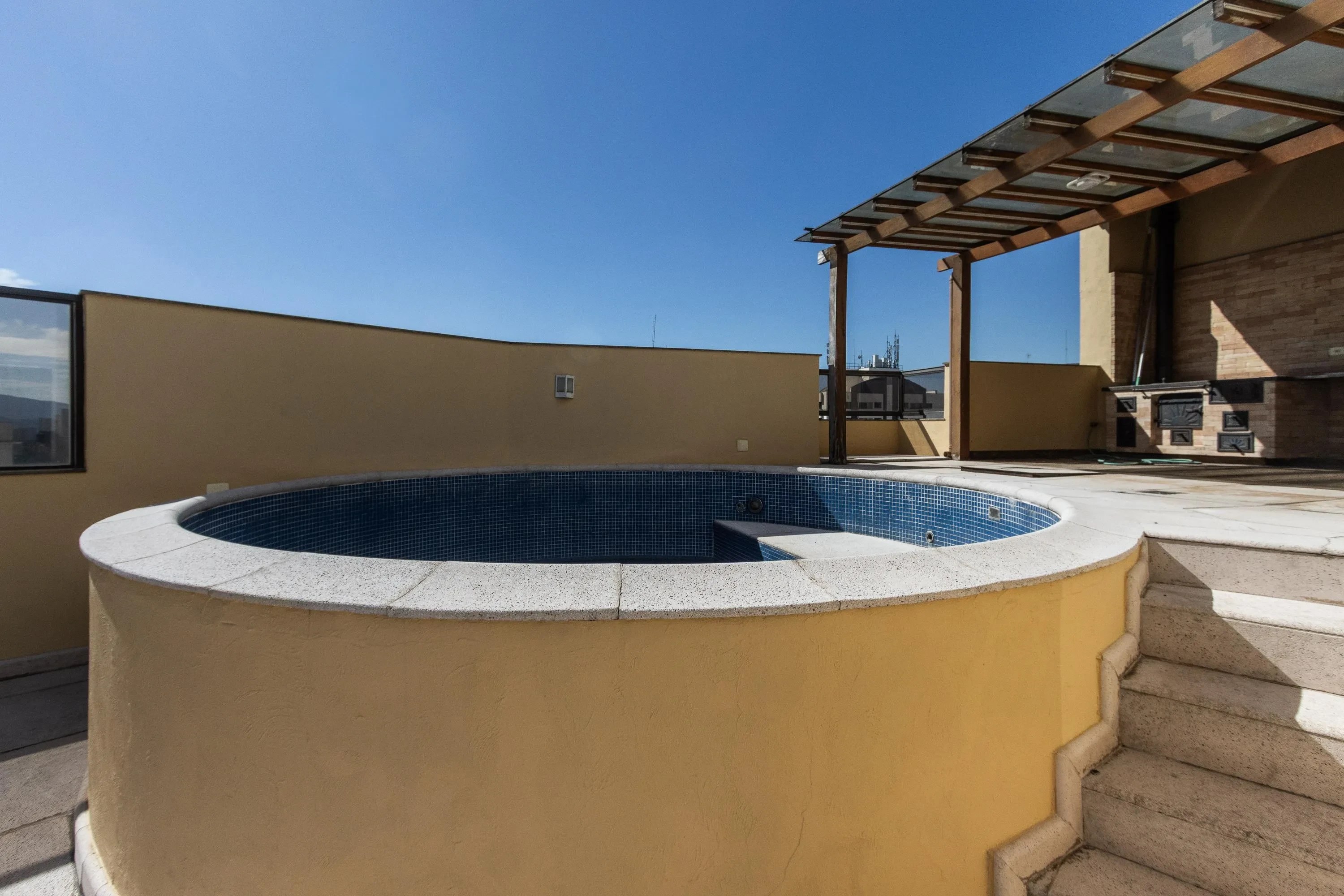 Piscina redonda de azulejos azuis em um terraço com paredes amarelas, degraus de mármore e um pergolado de madeira e vidro. Ao fundo, uma churrasqueira de tijolos e o céu azul claro.