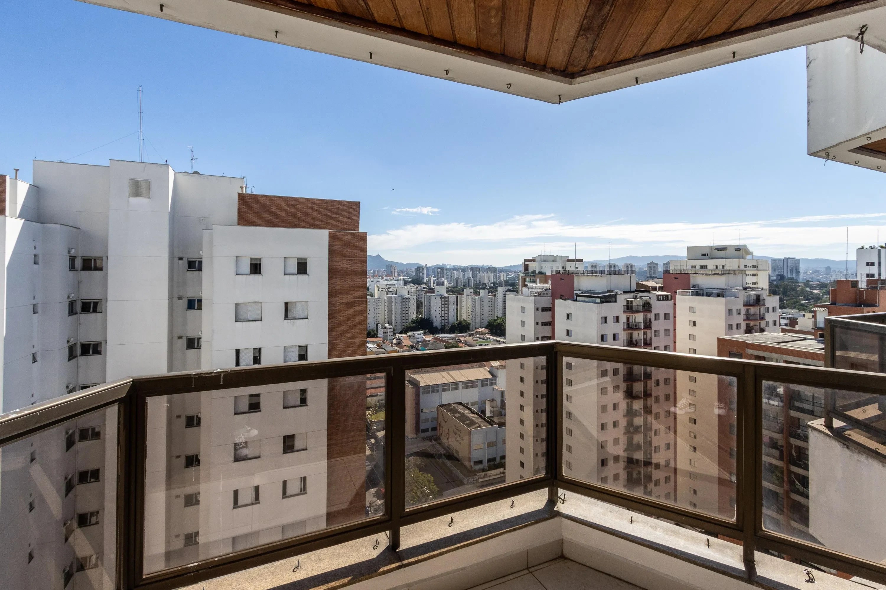 Vista de uma varanda com guarda-corpo de vidro e metal, mostrando prédios residenciais brancos e marrons sob um céu azul claro. Ao fundo, uma cidade com montanhas distantes e poucas nuvens esparsas