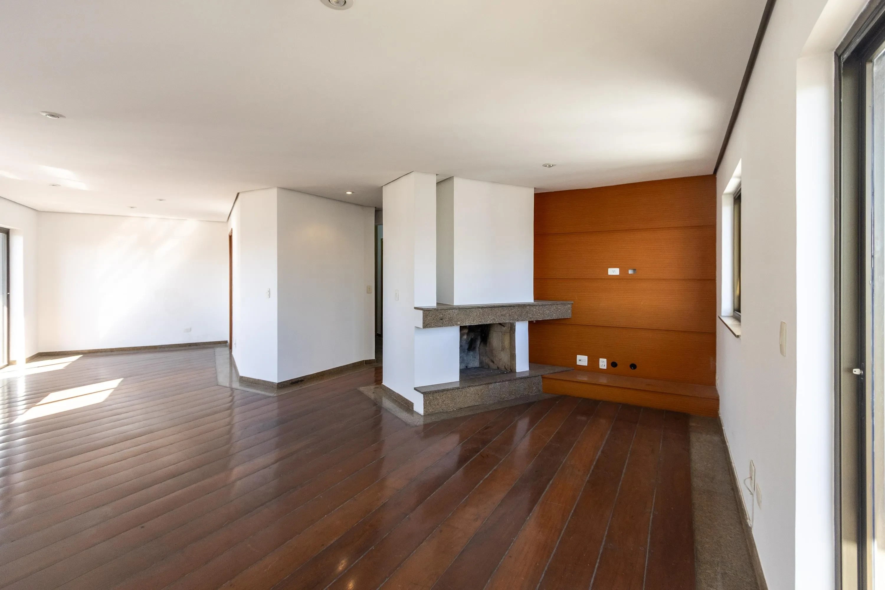 Sala de estar ampla e vazia com piso de madeira brilhante, paredes brancas e uma lareira central de pedra cinza. Uma parede ao fundo é revestida em madeira marrom-avermelhada, com tom quente. A luz do sol entra pela janela à esquerda, criando reflexos no chão.
