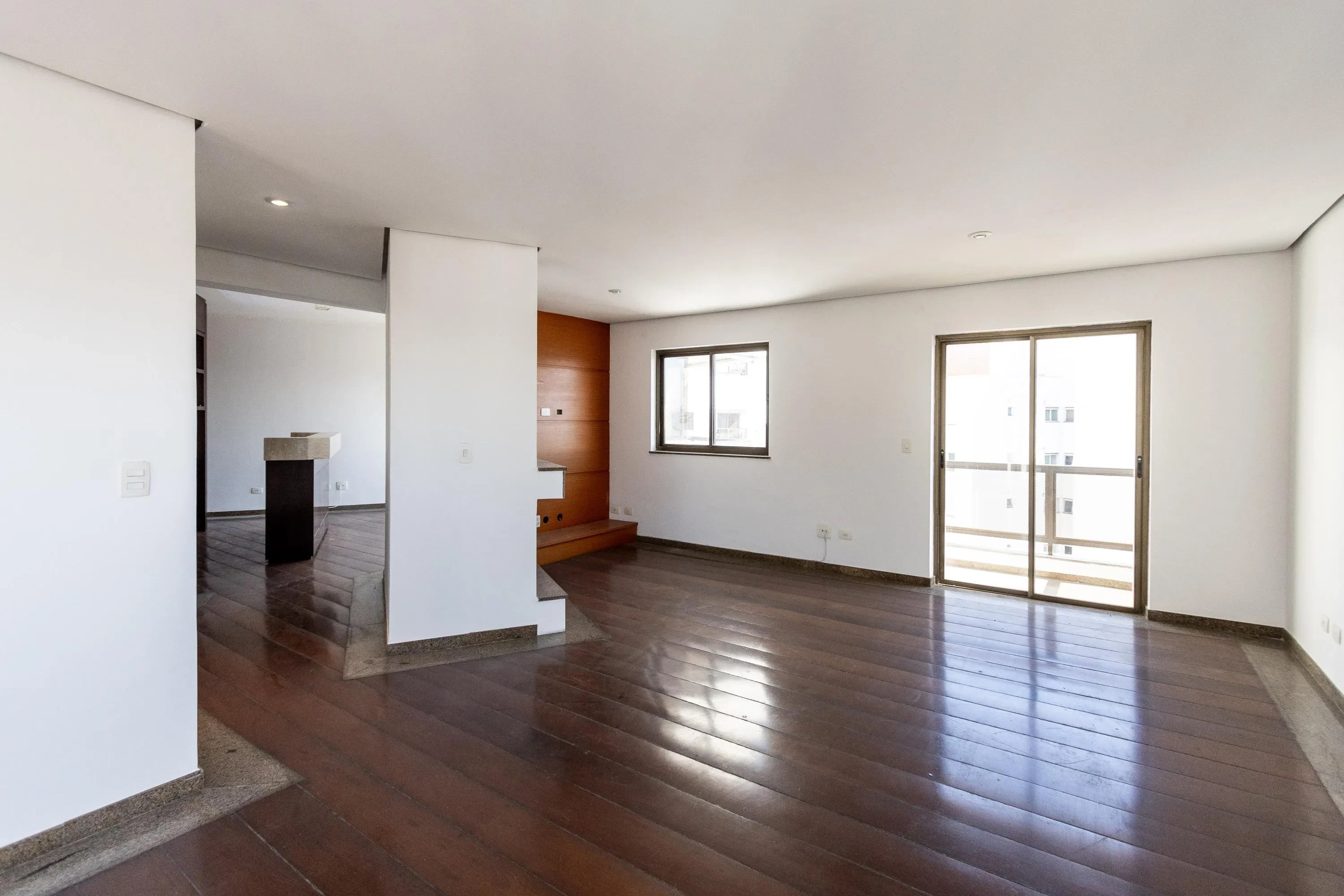 Sala ampla e vazia com paredes brancas e piso de madeira escura brilhante. À direita, uma porta de vidro deslizante dá para uma varanda, e uma janela retangular acima de um balcão de madeira. À esquerda, um corredor com um balcão escuro no centro. A iluminação natural entra pelas aberturas, criando reflexos no chão.