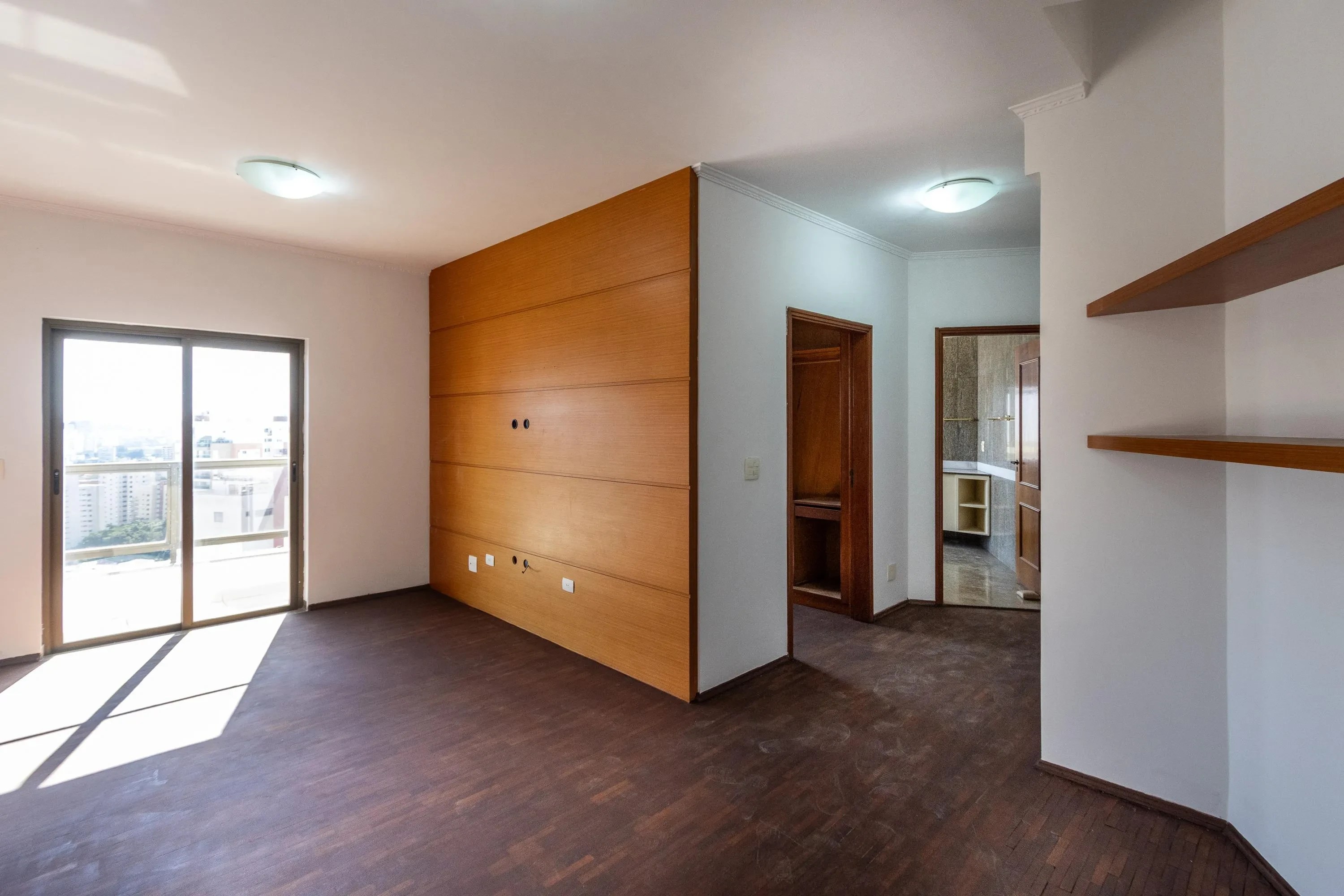 Sala vazia com piso de madeira escura, paredes brancas e uma parede lateral revestida em madeira clara. Há uma porta de correr de vidro que dá para uma varanda, e duas portas internas abertas revelando outros cômodos. Prateleiras de madeira clara estão visíveis na parede direita