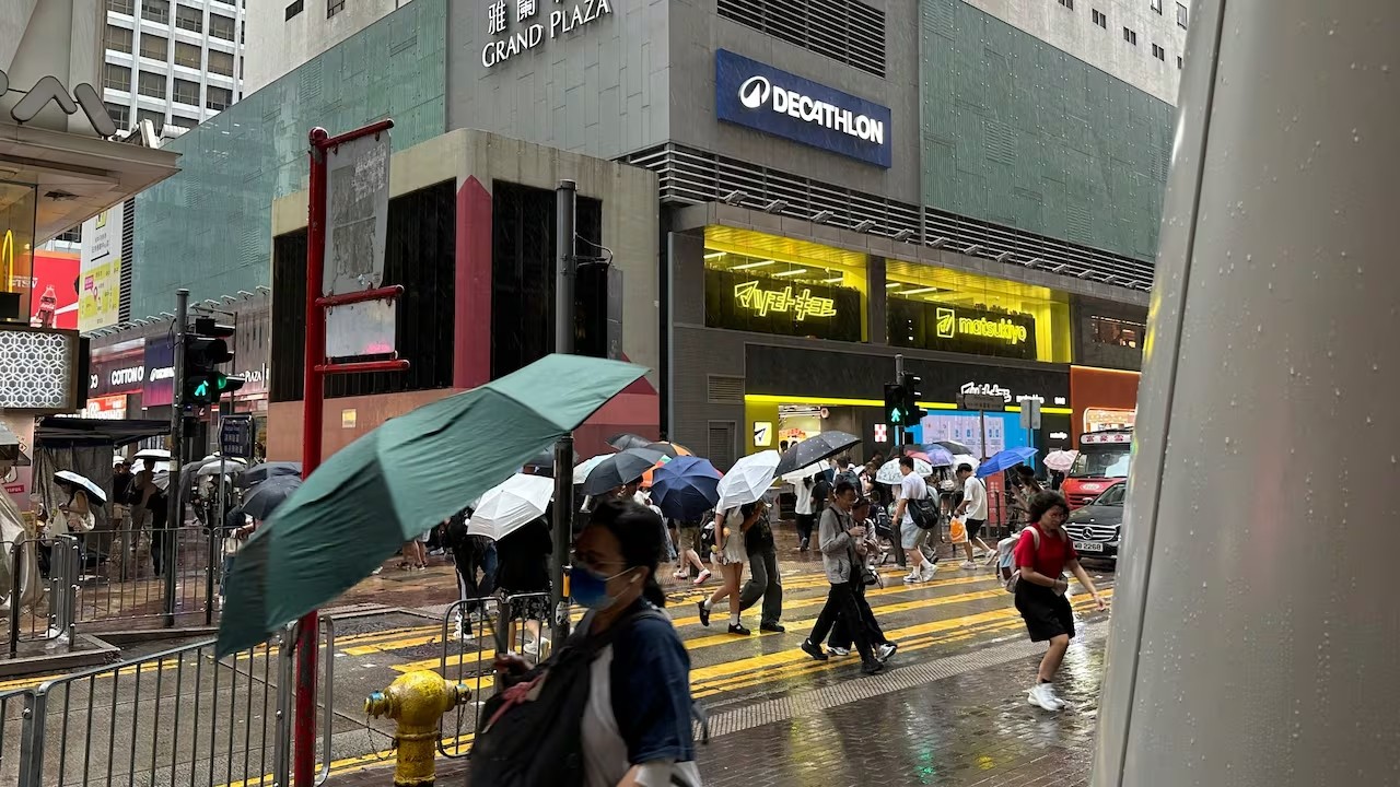 Pessoas com guarda-chuvas coloridos atravessam uma rua molhada em uma cidade asiática, com prédios altos e letreiros luminosos ao fundo, sob chuva