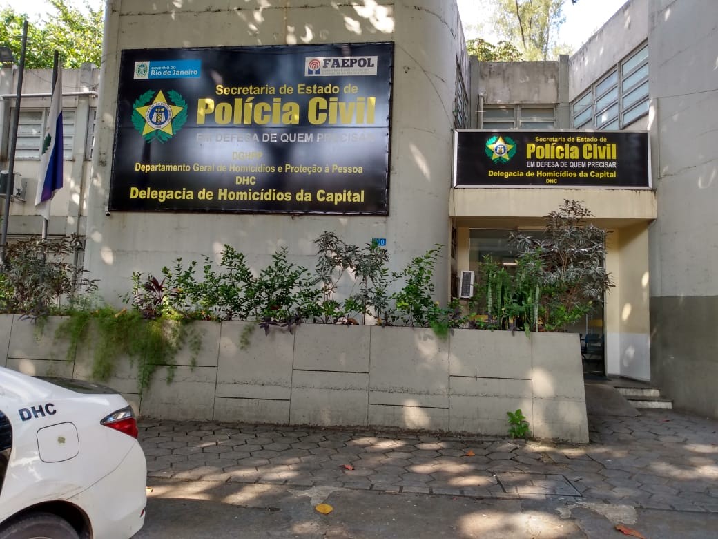Fachada da Delegacia de Homicídios da Capital (DHC) no Rio de Janeiro, com dois letreiros pretos e amarelos da Polícia Civil. Um carro branco com DHC na lateral está estacionado à esquerda, e há plantas verdes em canteiros de concreto na frente do prédio. O sol projeta sombras no chão de paralelepípedos