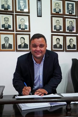 Homem de pele clara, camisa azul e blazer escuro, sorrindo enquanto assina documentos em uma mesa de madeira. Ao fundo, uma parede branca com vários retratos emoldurados de homens em preto e branco