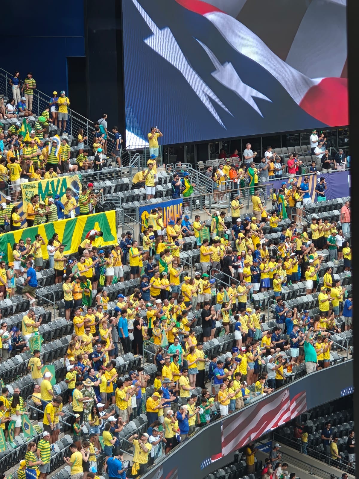 Multidão de torcedores em um estádio, a maioria vestindo camisas amarelas e algumas verdes, aplaudindo. Ao fundo, um telão exibe uma bandeira dos Estados Unidos e, em primeiro plano, uma faixa verde com a palavra MAVAs em amarelo