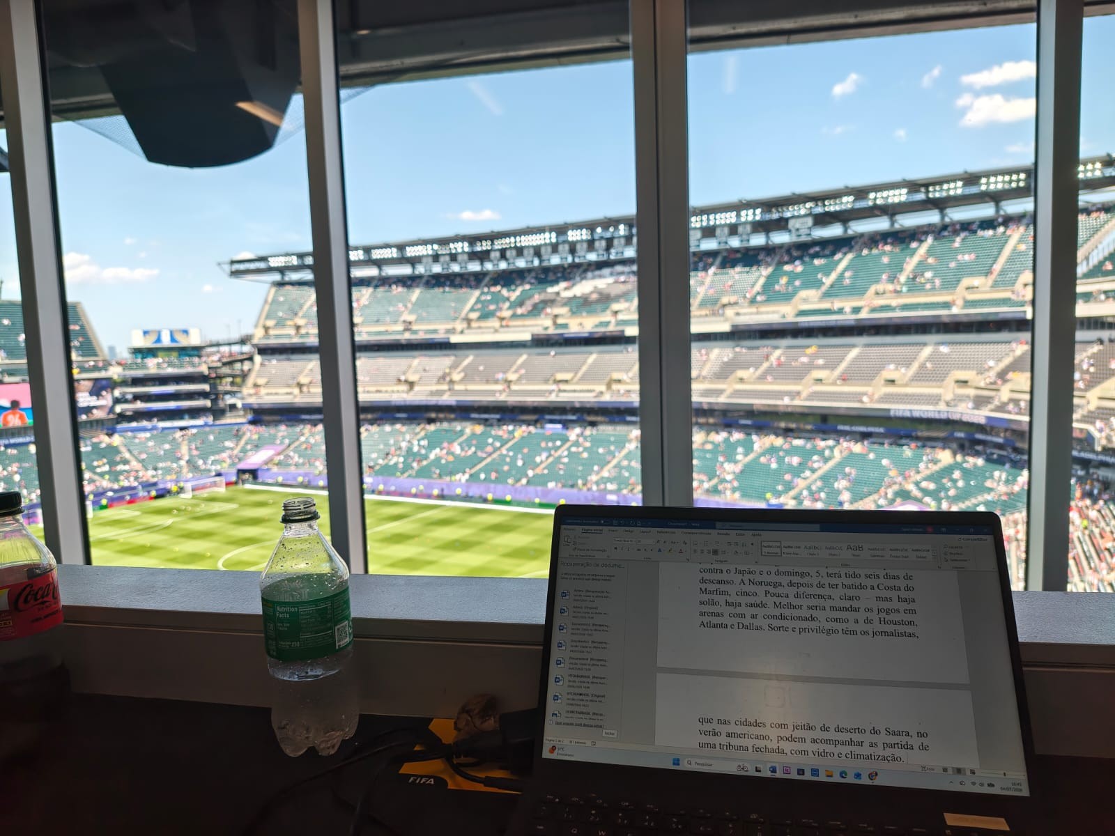 Visão de uma cabine de imprensa em um estádio de futebol, com um notebook aberto exibindo um texto e duas garrafas de bebida na bancada, enquanto o campo verde e as arquibancadas lotadas são visíveis através da janela