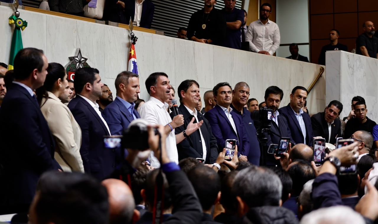 Políticos brasileiros, incluindo o ex-presidente Jair Bolsonaro, em um evento público, com alguns segurando celulares para gravar. Bolsonaro, de camisa branca, sorri enquanto fala ao microfone. Outros homens de terno e camisa social observam, alguns com expressões sérias. Bandeiras do Brasil e do estado de São Paulo são visíveis ao fundo, sugerindo um ambiente oficial. A cena é de um encontro político com grande presença de público e imprensa.