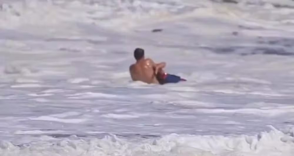 Homem de costas, sem camisa e com bermuda azul, sentado na água espumosa do mar, com uma prancha pequena vermelha ao lado