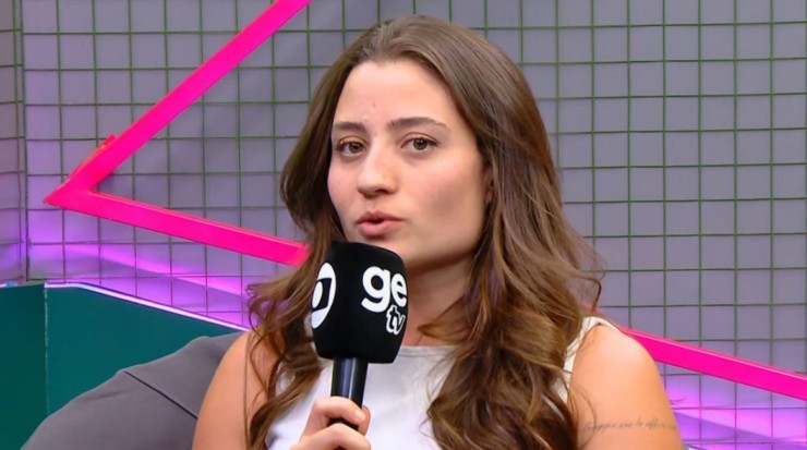 Jovem mulher de cabelos castanhos ondulados, vestindo blusa branca, segurando um microfone preto com o logo ge tv em branco, falando com a boca levemente aberta. Ao fundo, uma parede quadriculada com iluminação rosa e um objeto triangular rosa neon