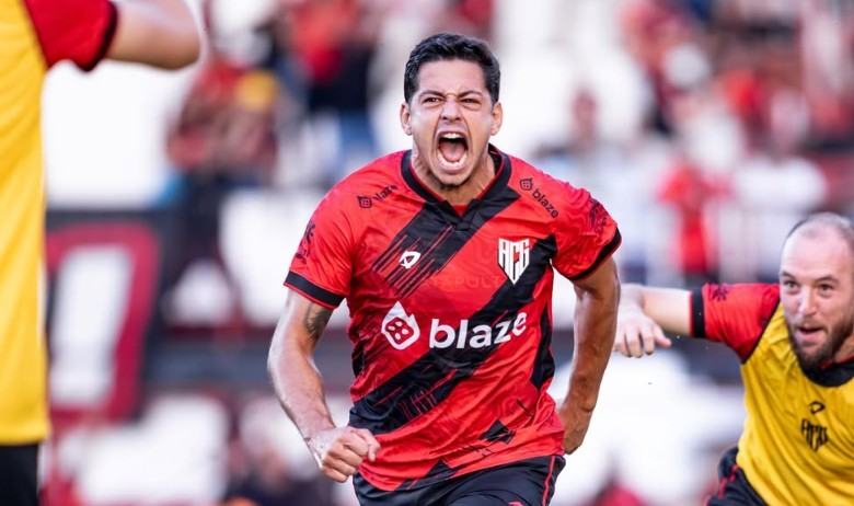 Jogador de futebol com camisa vermelha e preta, correndo e gritando com a boca aberta, em campo de futebol