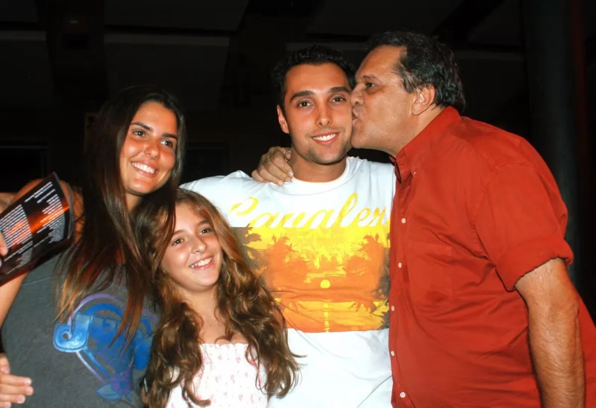 Quatro pessoas sorrindo para a câmera: duas mulheres jovens, um homem jovem e um homem mais velho que beija a bochecha do homem jovem