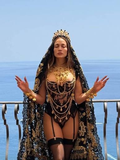 Jennifer Lopez em um body de renda preta com correntes douradas, coroa e véu bordado, braços abertos e olhos fechados, em uma varanda com vista para o mar azul