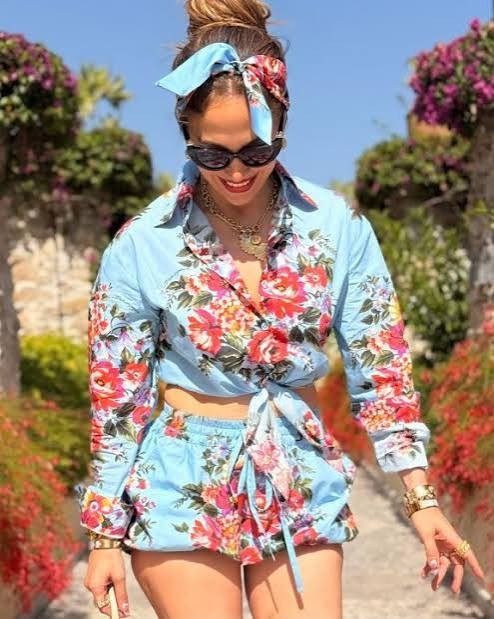 Mulher sorrindo, usando óculos escuros e um conjunto azul claro com estampa floral rosa e vermelha, blusa amarrada na cintura e shorts. Ela tem o cabelo preso em coque alto com um lenço combinando e usa colares dourados, pulseira e anéis. O fundo é um jardim ensolarado com folhagens e flores coloridas