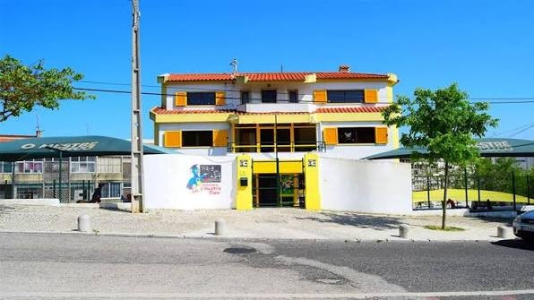 Fachada de um edifício de dois andares, branco com detalhes amarelos e telhado de telhas vermelhas, sob céu azul. A entrada principal é amarela, com um portão escuro. À esquerda, um muro branco exibe um desenho colorido de um pássaro azul e texto. À direita, uma árvore verde e um carro estacionado. Ao fundo, outras construções com telhados verdes. Um poste de luz cinza está à frente, no centro