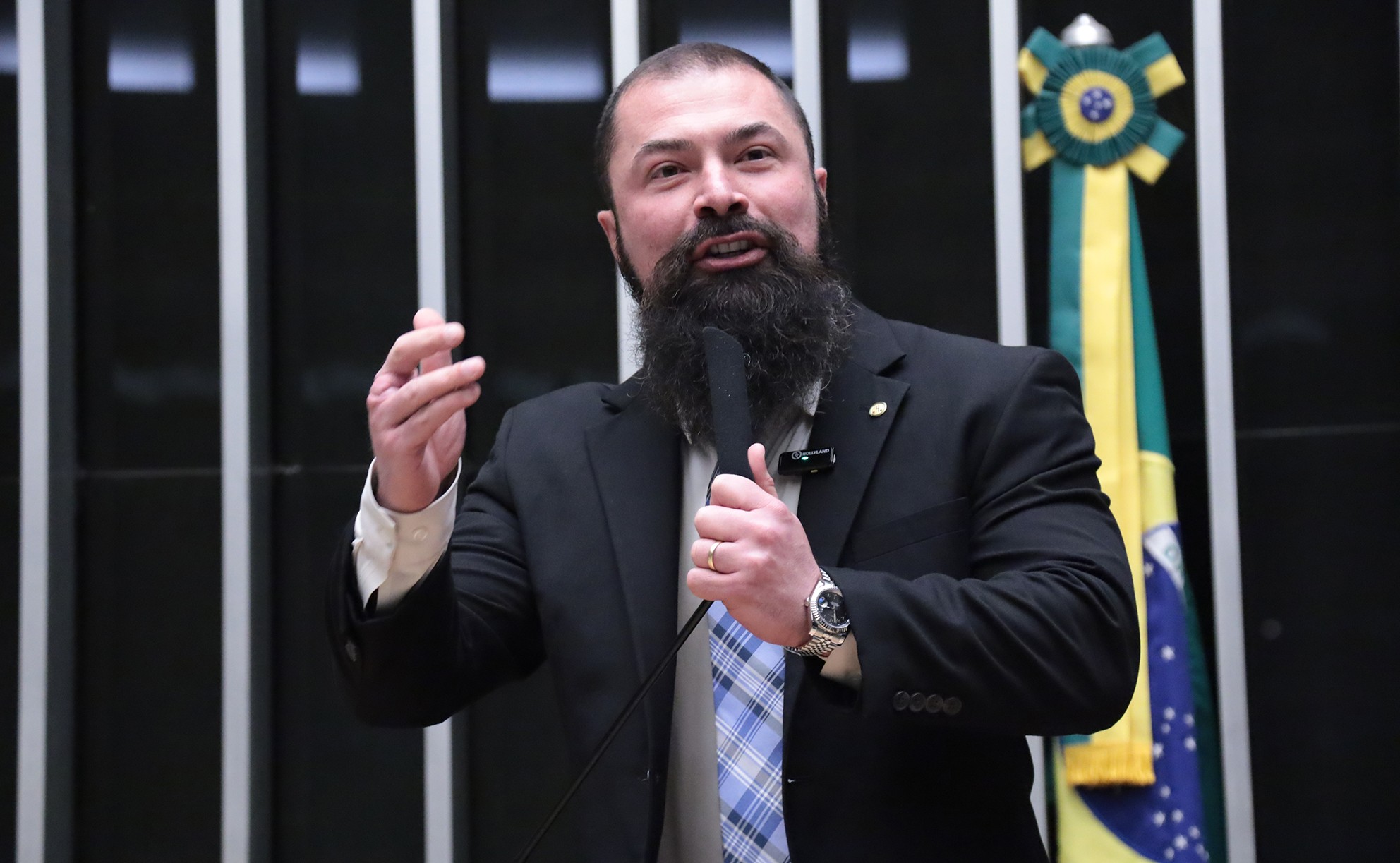 Homem barbudo, de terno preto e gravata xadrez azul, fala ao microfone com a mão esquerda levantada, em um ambiente formal com a bandeira do Brasil ao fundo