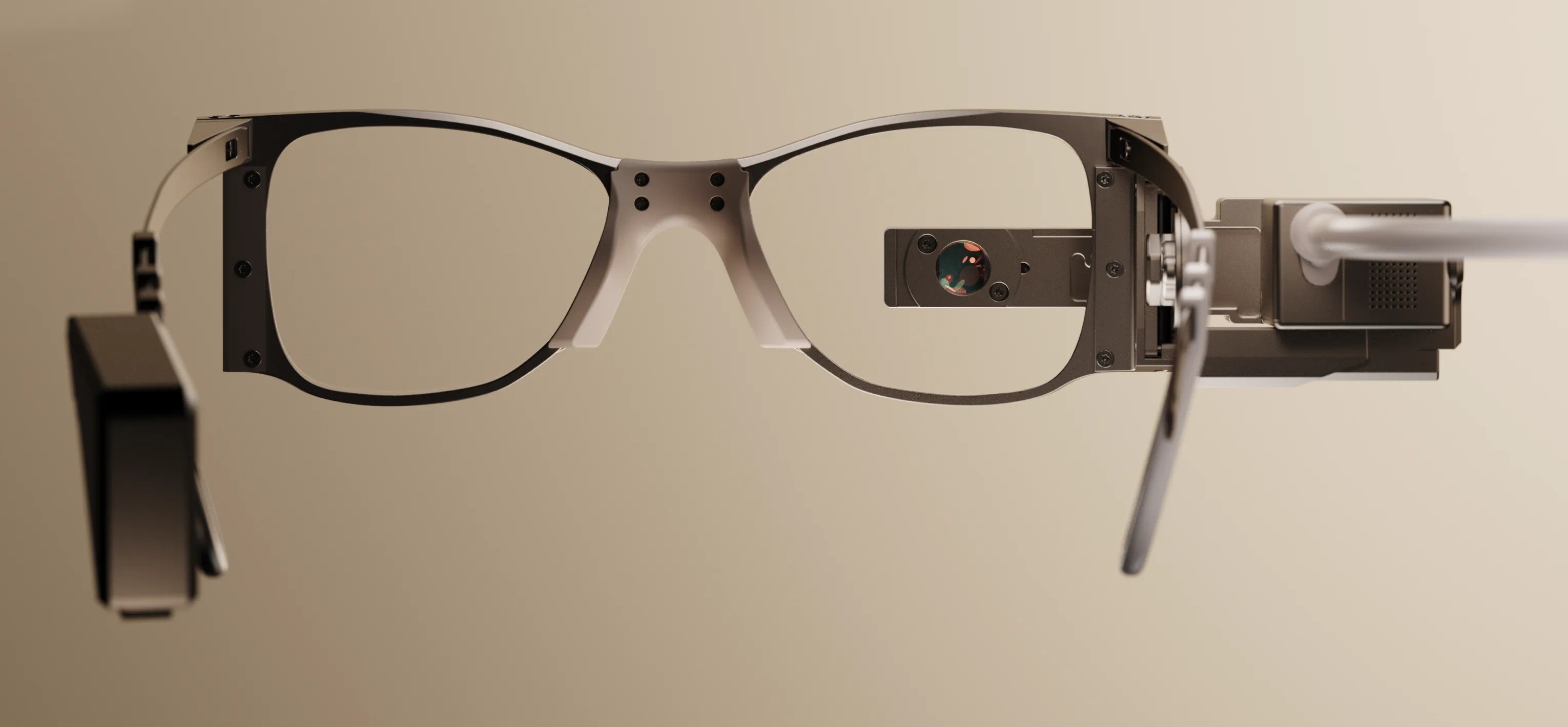 Óculos de realidade aumentada com armação cinza-escura e lentes transparentes, com uma pequena câmera acoplada à lente direita e um cabo branco conectado à haste direita, sobre um fundo bege