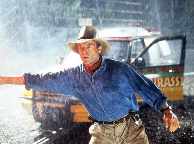 Homem de chapéu e camisa jeans azul, com lenço vermelho no pescoço, braço estendido para a esquerda, sob chuva forte, com um jipe do Jurassic Park ao fundo