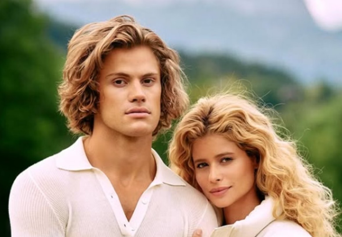 Um homem e uma mulher jovens, ambos loiros e de pele clara, posam juntos. Ele, à esquerda, tem cabelos ondulados e veste uma camisa polo branca. Ela, à direita, tem cabelos cacheados e apoia a cabeça no ombro dele, olhando para a frente. O fundo é desfocado, com tons verdes e azuis de natureza