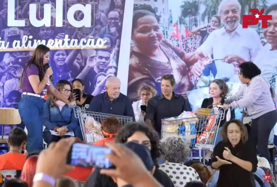 Mulher de blusa roxa e calça jeans fala ao microfone, enquanto Lula e Fernando Haddad, ambos de camisa escura, observam carrinhos de supermercado cheios de alimentos. Ao fundo, um banner gigante com a palavra Lula e alimentação, e fotos de pessoas sorrindo. Uma intérprete de Libras gesticula na frente, e pessoas na plateia filmam com celulares