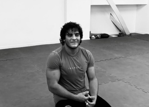 Homem jovem de cabelo escuro e encaracolado, vestindo camiseta de treino, sentado em tatame cinza, sorrindo levemente para a câmera em um ambiente de academia