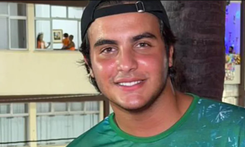 Homem jovem de pele clara, cabelo escuro e boné preto virado para trás, sorrindo para a câmera. Veste uma camiseta verde com estampa de folhas. Ao fundo, uma janela com pessoas e um coqueiro