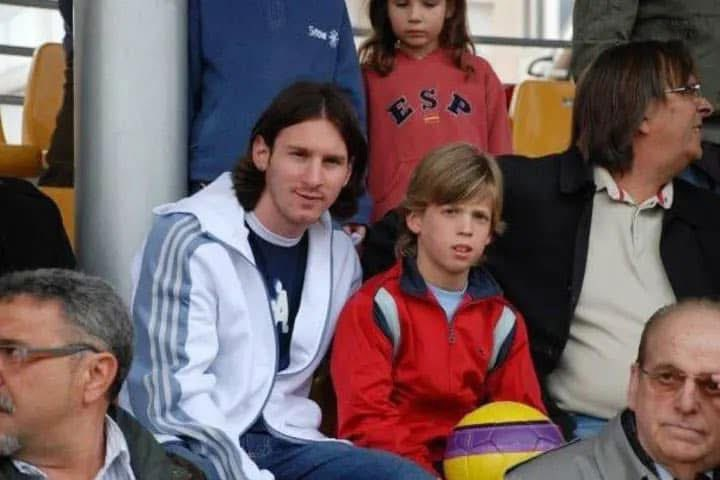 Lionel Messi jovem, com cabelo comprido, sorri levemente ao lado de um menino loiro de jaqueta vermelha, segurando uma bola colorida. Ambos estão sentados em uma arquibancada, com outras pessoas ao redor