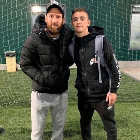 Lionel Messi, com barba e boné, usando jaqueta preta e calça cinza, abraça um jovem de moletom preto e mochila cinza, em campo de futebol com rede verde ao fundo