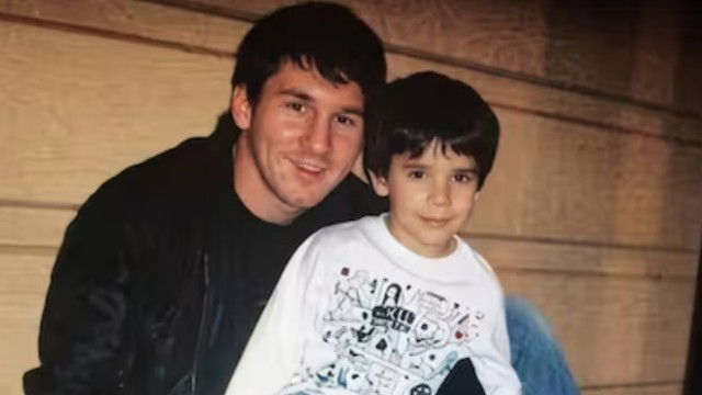 Lionel Messi jovem, com cabelo escuro e jaqueta preta, sorri abraçando um menino pequeno de cabelo escuro e camiseta branca estampada, que olha para frente com expressão séria