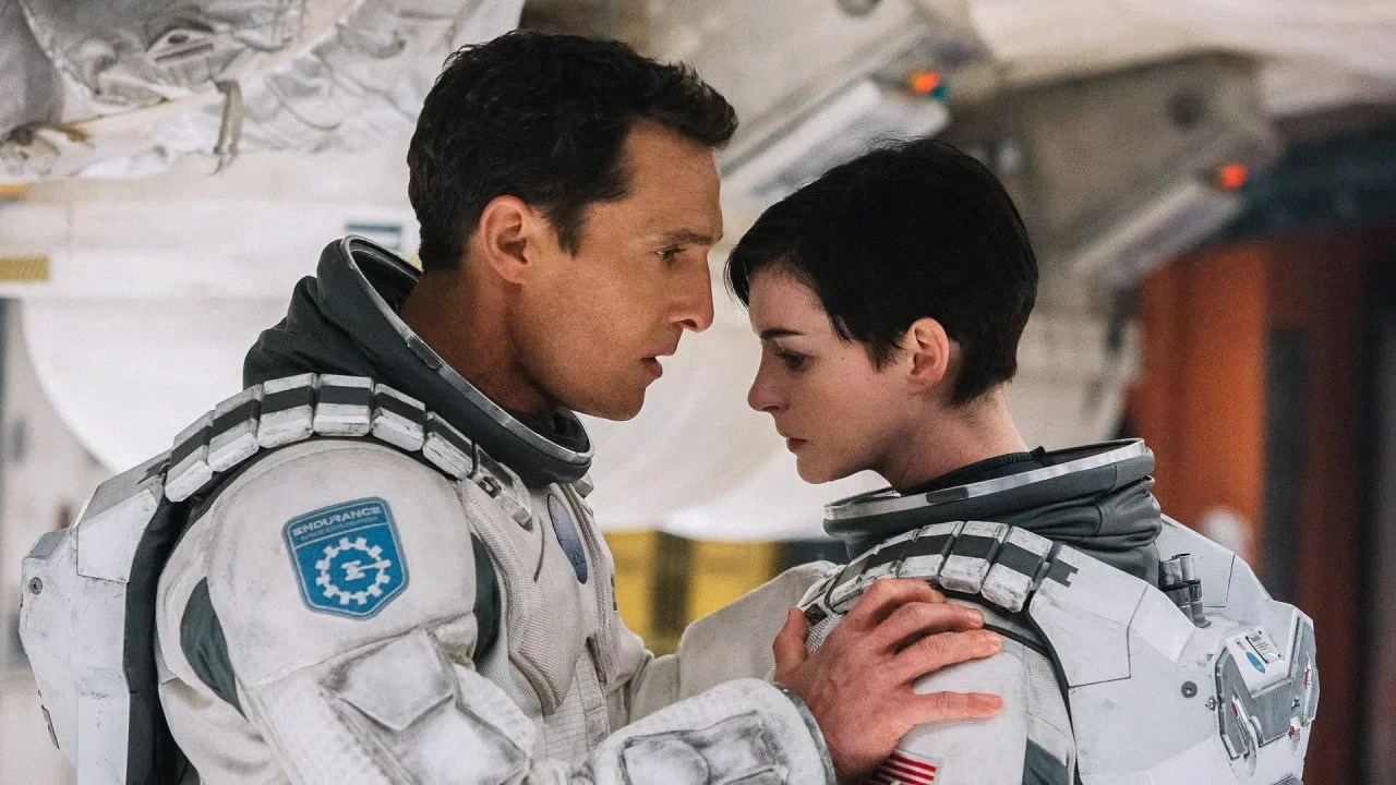 Um homem e uma mulher, ambos em trajes espaciais brancos, se olham intensamente em um ambiente de nave espacial. O homem, à esquerda, tem cabelos castanhos e expressão séria. A mulher, à direita, tem cabelos curtos e escuros, com a mão do homem em seu ombro. Ambos usam emblemas da missão Endurance em seus ombros.