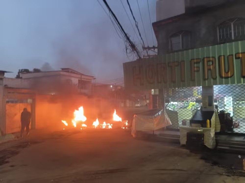 Chamas intensas e fumaça escura em uma rua, com um homem de costas à esquerda e um estabelecimento HORTIFRUTI com portas fechadas à direita