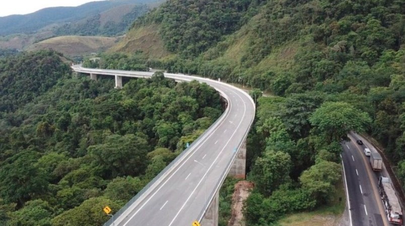 Vista aérea de uma rodovia sinuosa com viadutos e pontes, cortando uma densa floresta verde em uma área montanhosa. Um caminhão e outros veículos trafegam na pista inferior, enquanto a pista superior está vazia. O céu está nublado, indicando um dia cinzento