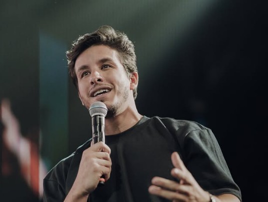 Homem jovem, branco, cabelo castanho e barba rala, sorrindo e olhando para cima, segurando um microfone com a mão direita e gesticulando com a esquerda, vestindo camiseta preta, em palco escuro com iluminação focada