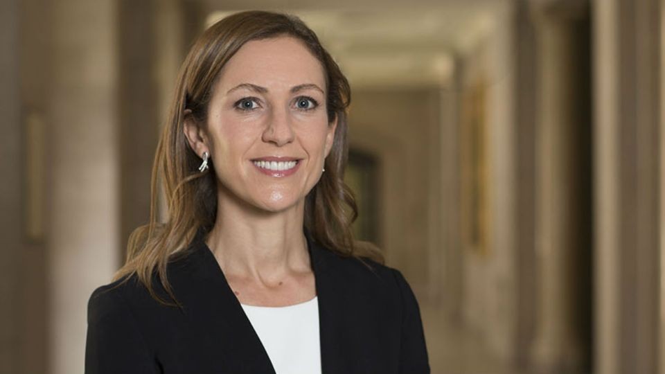 Mulher de cabelos castanhos e compridos, sorrindo, vestindo blazer preto e blusa branca, em um corredor com paredes claras e colunas