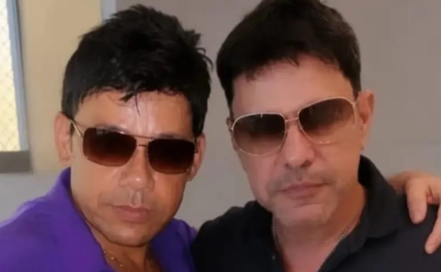Dois homens de pele clara, com cabelos escuros e óculos de sol, posam lado a lado. O da esquerda veste uma camisa roxa e o da direita, uma camisa preta, com a mão no ombro do outro