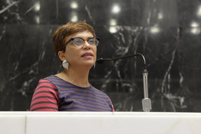 Mulher de pele clara, cabelo curto castanho, óculos de armação preta e branca, e brincos prateados em formato de folha, fala em um microfone. Ela veste uma blusa listrada em tons de azul e vermelho, e está em um ambiente com parede escura de mármore e uma bancada branca à frente