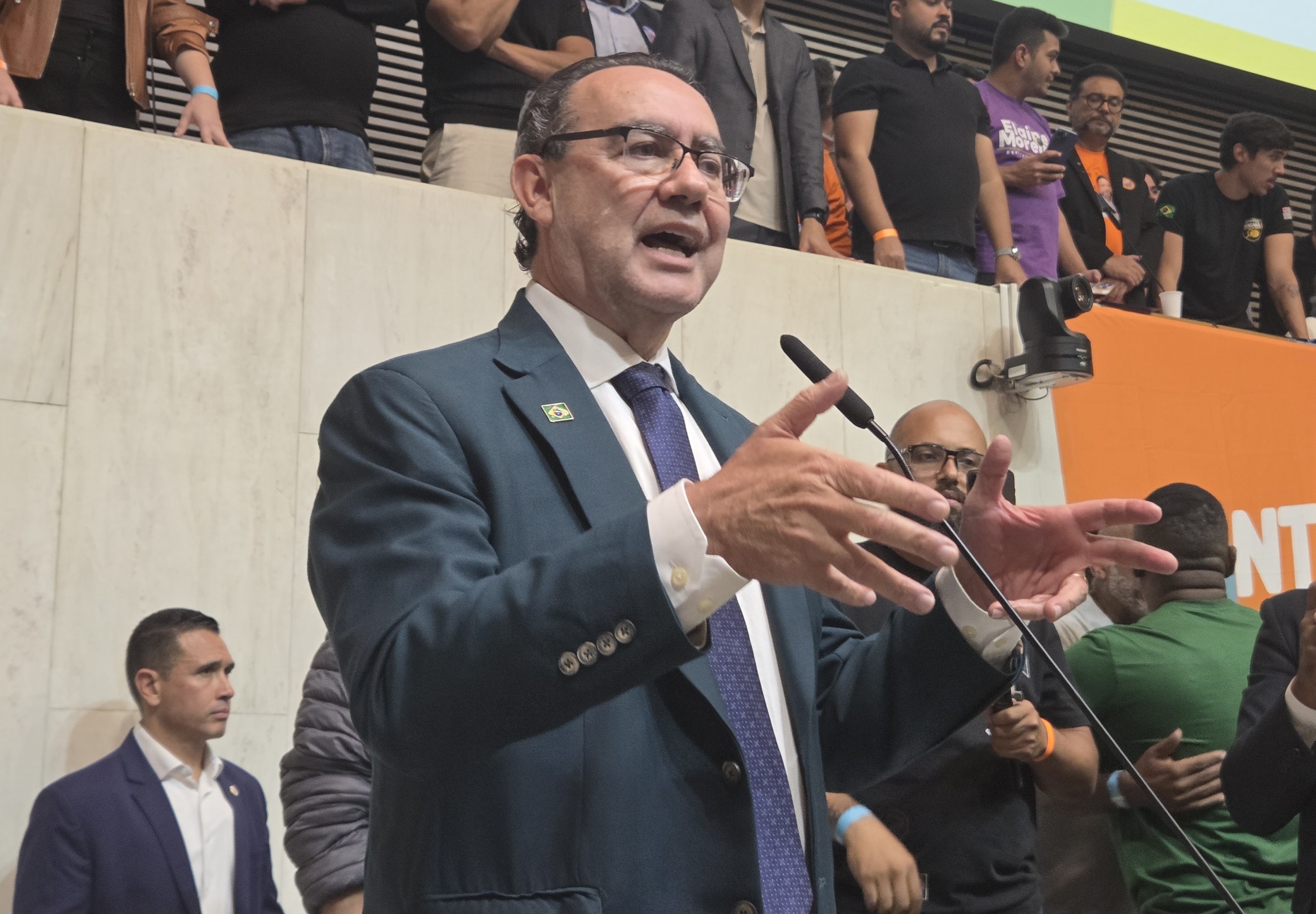 Homem de terno azul-marinho, camisa branca e gravata azul, com óculos, fala em um microfone, gesticulando com as mãos abertas. Ao fundo, pessoas assistem em um auditório, com uma faixa laranja visível à direita