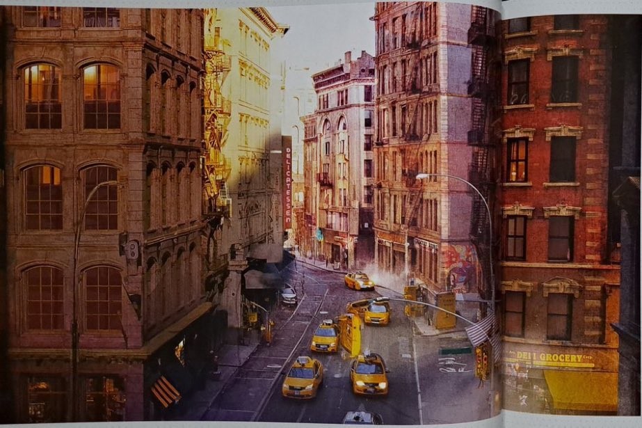 Rua movimentada em Nova York ao entardecer, com pr&eacute;dios antigos de tijolos e janelas iluminadas. V&aacute;rios t&aacute;xis amarelos circulam pela via, e bandeiras americanas decoram alguns postes. A luz dourada do sol poente reflete nas fachadas, criando um contraste de cores e sombras. Um letreiro de DELI GROCERY &eacute; vis&iacute;vel em um dos edif&iacute;cios.