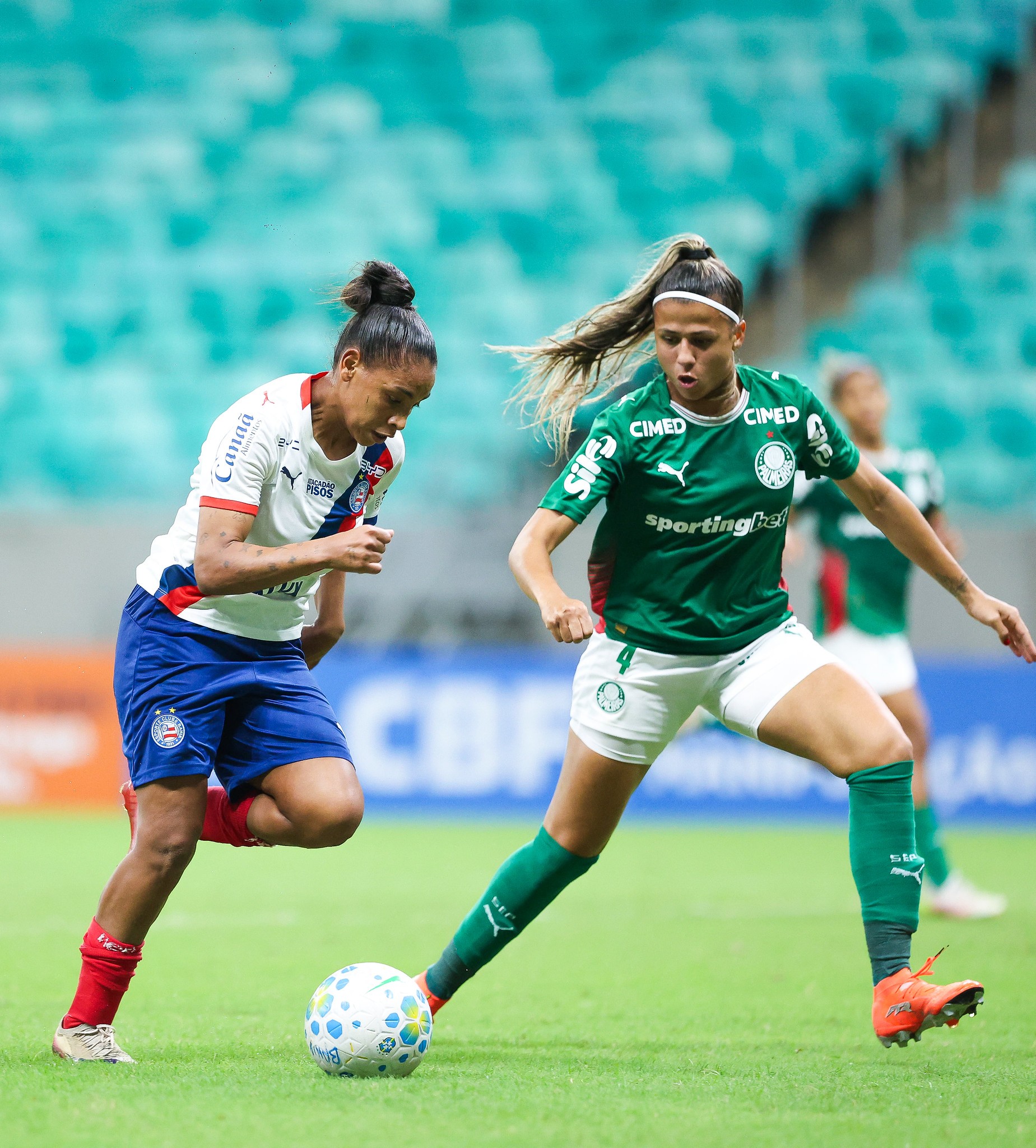 Duas jogadoras de futebol em campo verde. Uma, &agrave; esquerda, veste camisa branca, shorts azul e meias vermelhas, com cabelo preso. A outra, &agrave; direita, usa camisa verde, shorts branco e meias verdes, com cabelo preso em rabo de cavalo. A jogadora de verde est&aacute; com o p&eacute; na bola branca e azul, enquanto a de branco a observa, ambas em movimento durante a partida. Ao fundo, arquibancadas azuis desfocadas