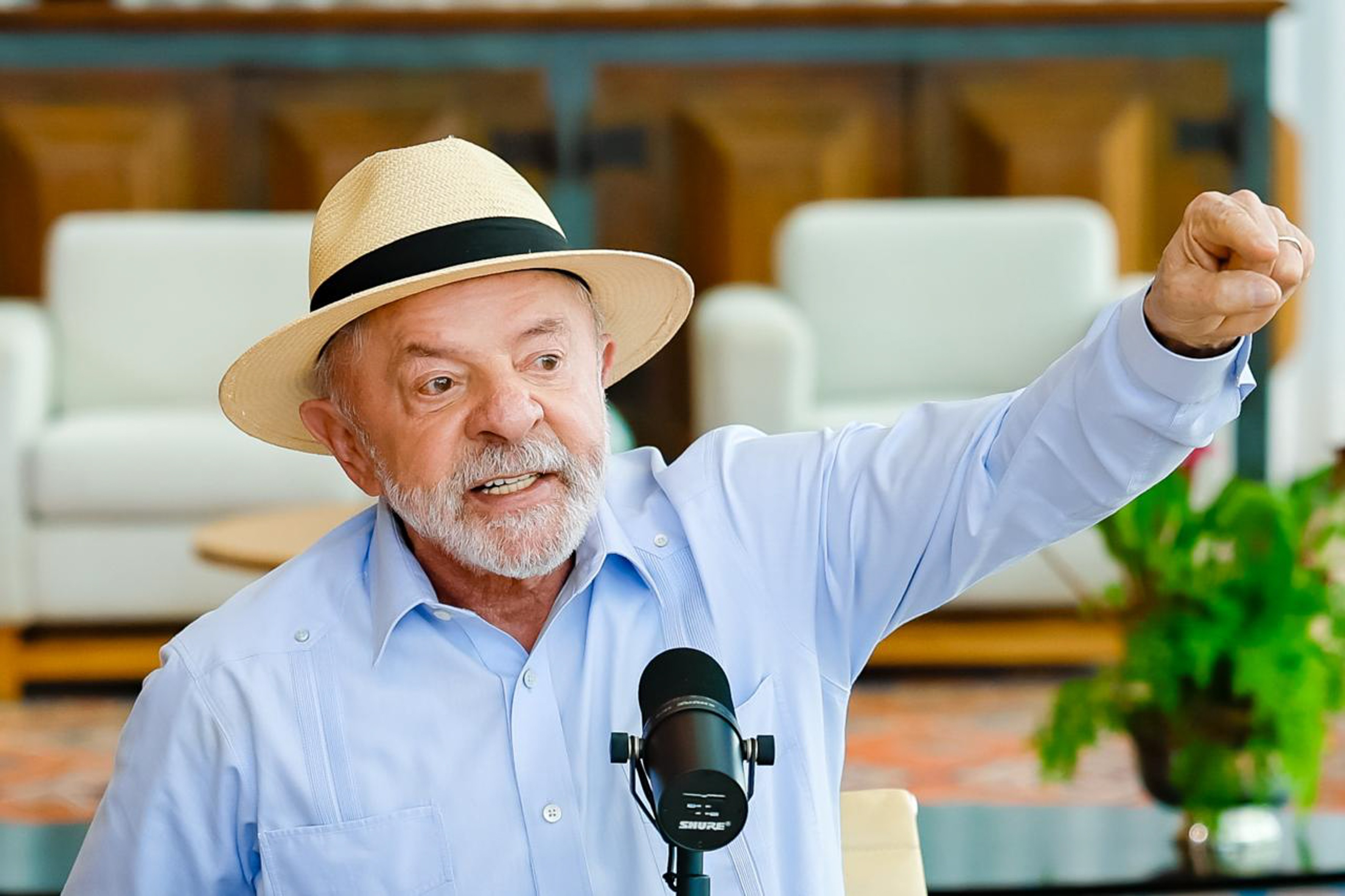 Homem idoso, de barba branca e chap&eacute;u de palha, vestindo camisa azul clara, gesticula com o bra&ccedil;o direito erguido e punho cerrado, falando em um microfone preto. Ao fundo, m&oacute;veis claros e uma planta verde