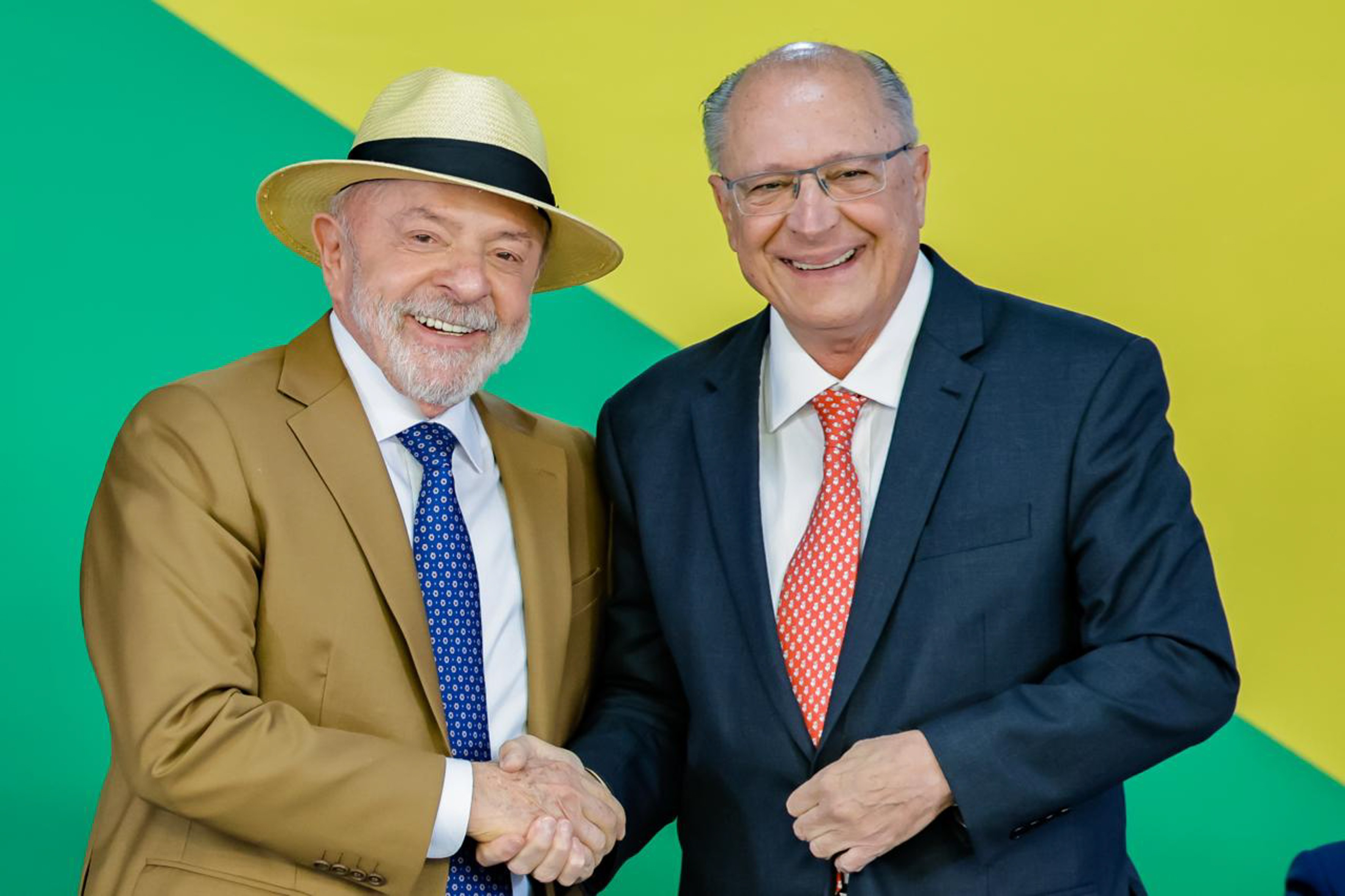Lula, de chapéu de palha e terno marrom, e Geraldo Alckmin, de terno azul-marinho e óculos, sorriem enquanto apertam as mãos, com fundo verde e amarelo