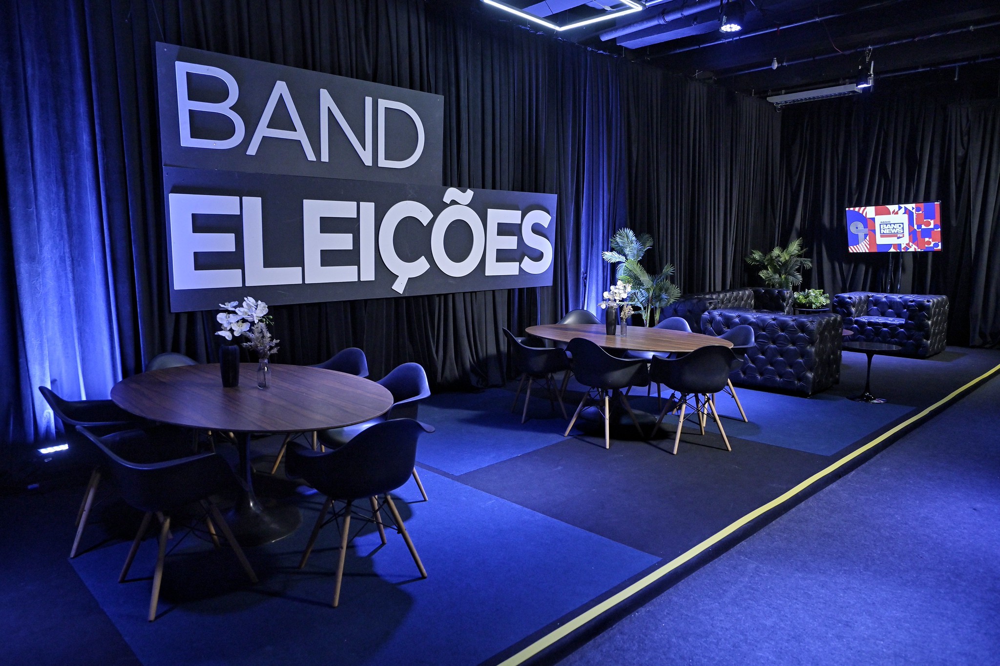 Ambiente de estúdio com cortinas pretas e iluminação azul, exibindo um banner BAND ELEIÇÕES. Há mesas redondas de madeira com cadeiras pretas, sofás azuis escuros e plantas decorativas. Uma televisão exibe uma imagem colorida no fundo